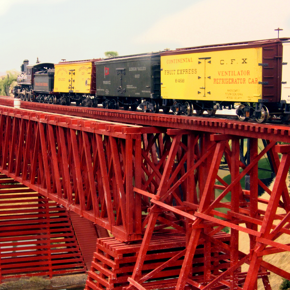 Color photo of steam locomotive and train passing over wood trestle and truss bridge.