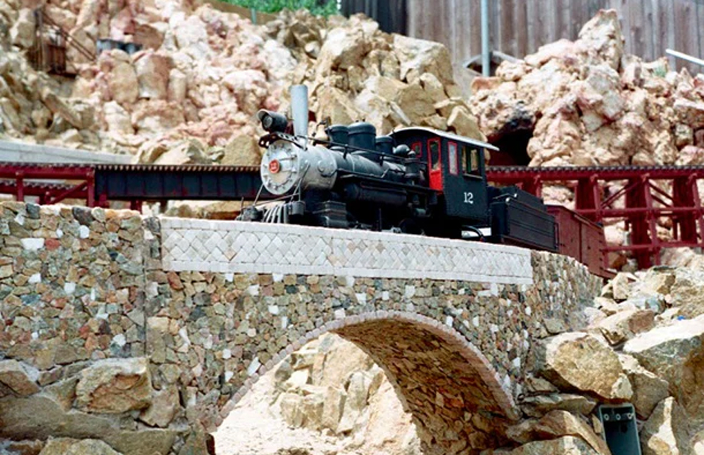 A model train led by a steam locomotive crosses a bridge on an outdoor garden railroad
