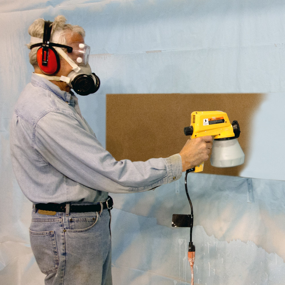 Color photo of man in denim shirt and blue jeans wearing personal protective gear painting tempered hardboard.