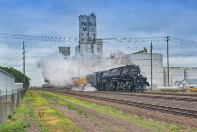 Big Boy steam locomotive and train passing a grain elevator in a rural town.