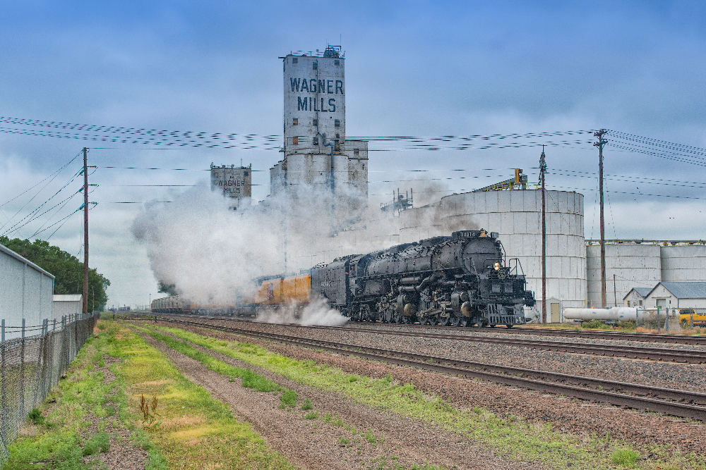 Big Boy steam locomotive and train passing a grain elevator in a rural town.