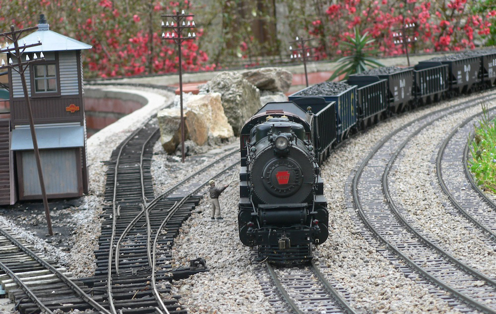 A scale train on an outdoor train layout with a trackside figure and structure