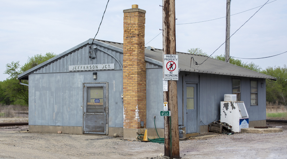 Color photo of gray metal building with doors, windows, and brick chimney.