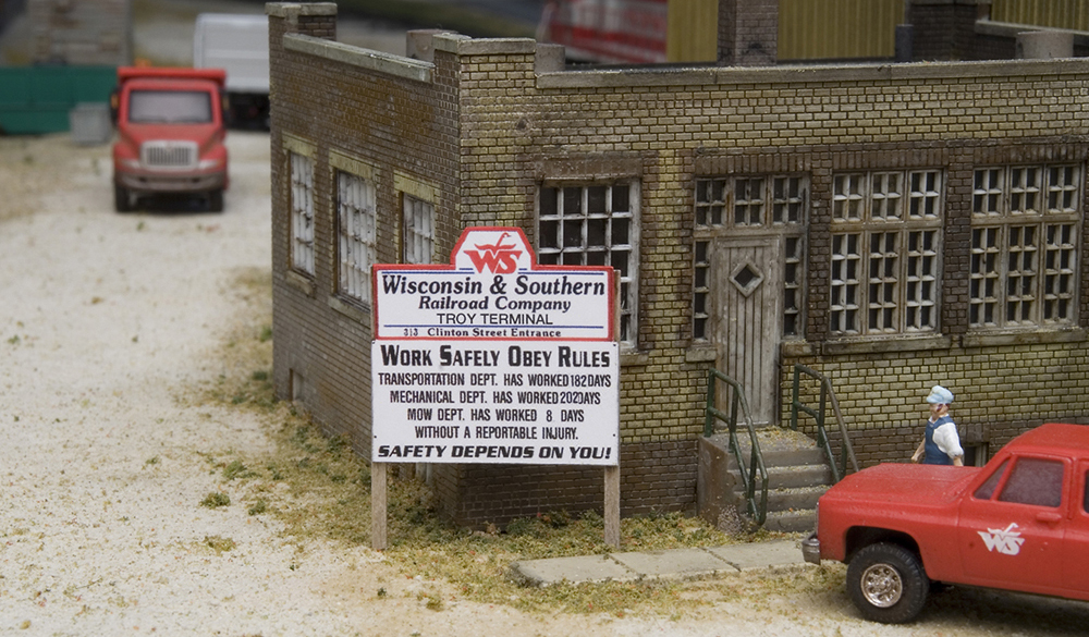 A railroad sign on a model railroad layout next to an industrial structure with a red truck and a figure seen nearby