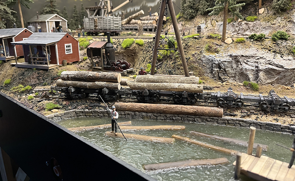 Highly detailed model railroad scene showing a pond with green water surrounded by gray rocks and tan dirt with green trees and red and white buildings.