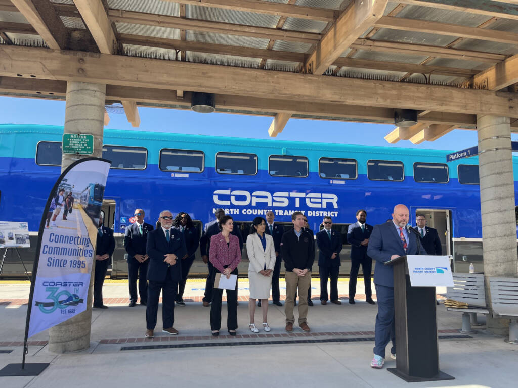 Man speaking at podium on station platform with blue railcar in background