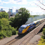 Blue, yellow, red, and white locomotive leads train of bilevel commuter cars with city skyline in background