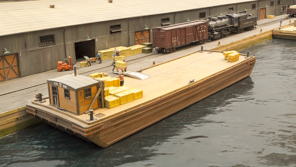 A barge in the foreground of the image along a rail marine model railroad layout, with a steam locomotive and freight car in the background