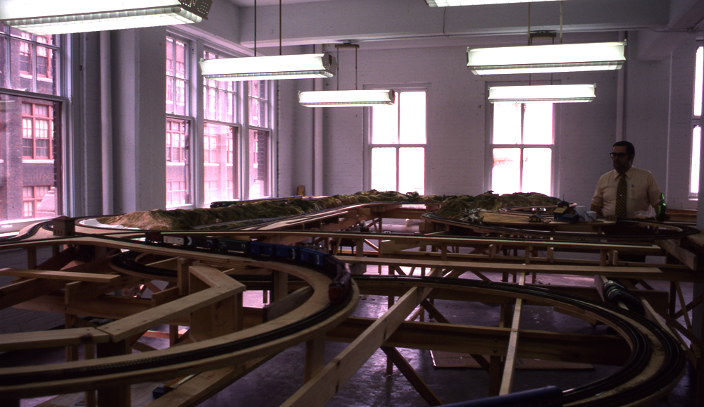 Remembering original Milwaukee Racine Troy: A color photograph of a partially completed, partially scenicked model railroad layout, with a man operating a model train on the layout