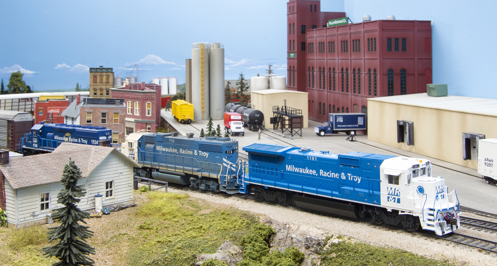 new era Milwaukee Racine Troy: A model train led by three blue and white locomotives proceeds through a town