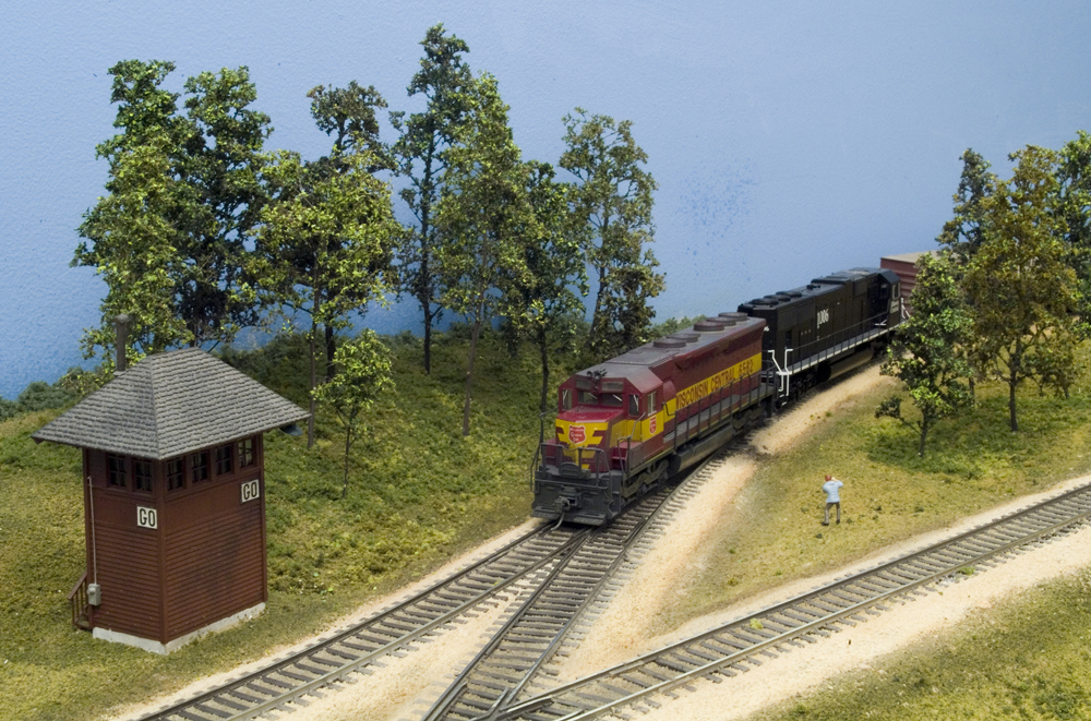 Color photo of two diesels passing a tower and trees.