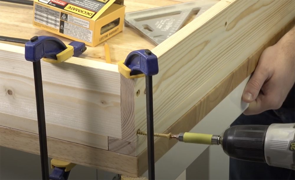 Various tan boards are clamped to a workbench with blue, yellow, and black clamps, forming a box. Also in the picture is a drill with a screwdriver bit turning a yellow screw into a small hole in the boards.