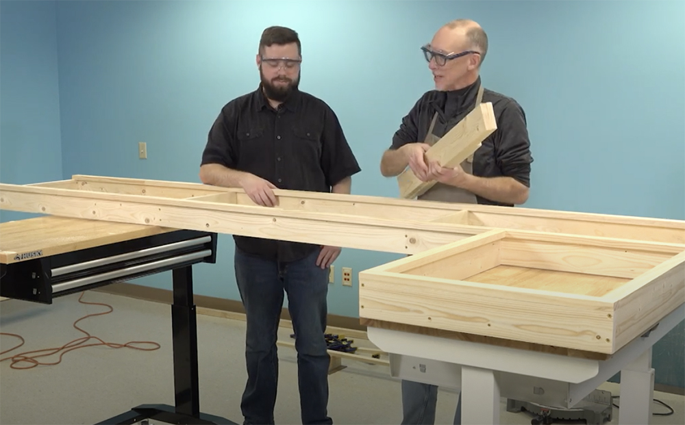 bearded young man in a dark shirt holds a rectangular frame made of bare wood boards in his right hand while a second, older man with thinning gray and wearing a tan apron hair carries several smaller boards in both hands.