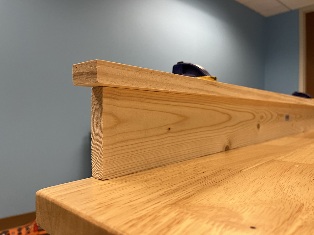 A tan board with a smaller tan board on top forming an upside-down L shape all held to a wood workbench with a black and yellow clamp.