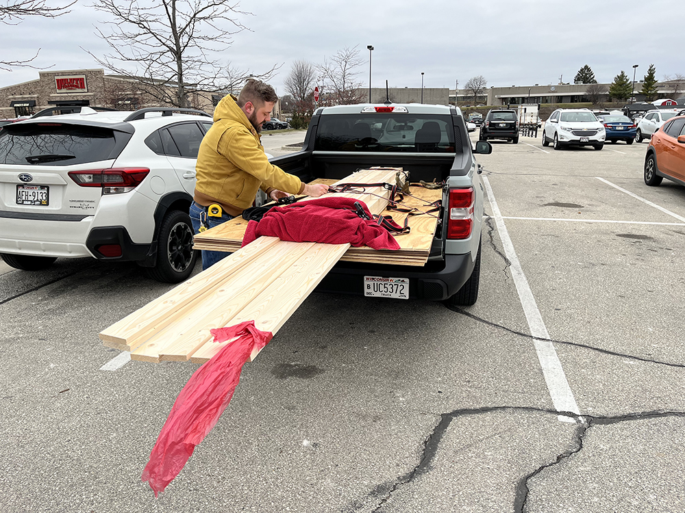Man in tan coat tying rope across a stack of new lumber loaded in the back of a gray pickup truck on a cloudy day.