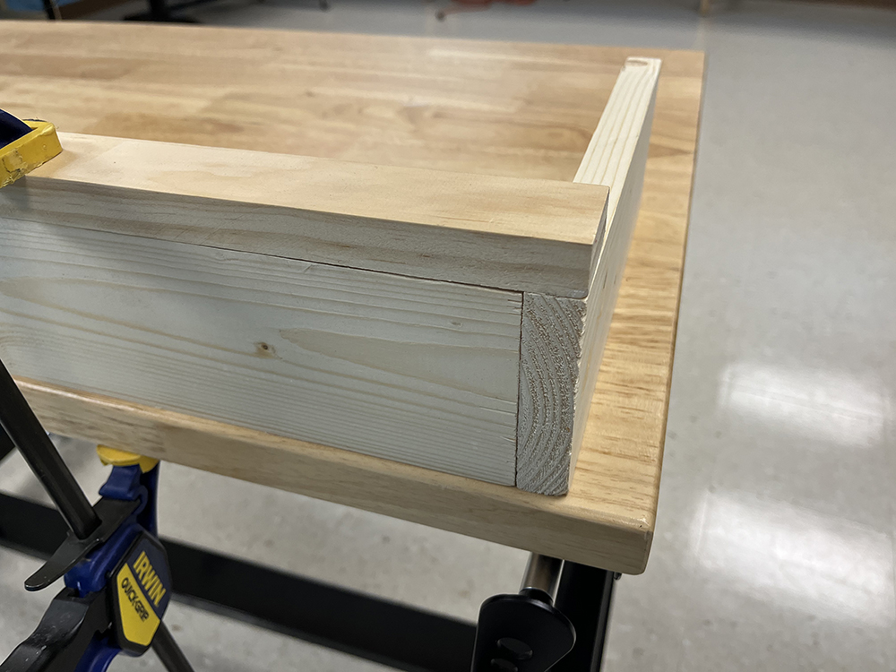 Three brown, unpainted boards held to a tan workbench with a blue and yellow bar clamp and featuring the corner where the board all connect with each other.