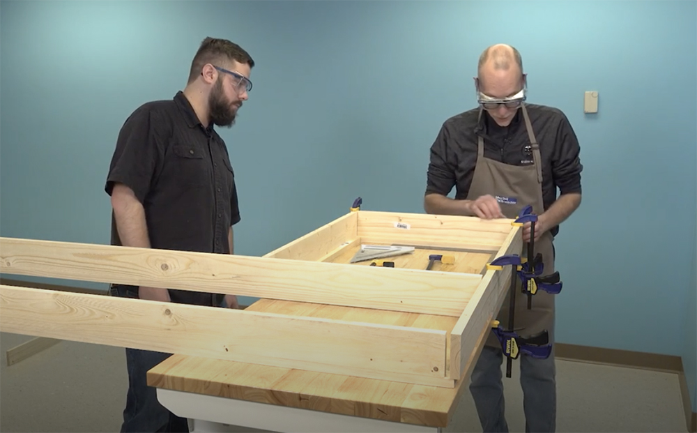 two men, one in a dark shirt and full beard and one in gray shirt and tan apron with a balding head, place tan boards on a wooden workbench to form two rectangles.