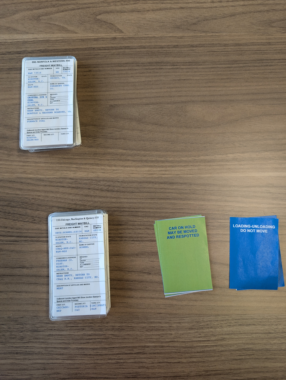 Model railroad car cards along with blue and green car tags on a wood table. These are used to operate the layout as a card game