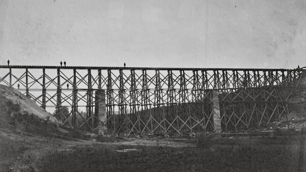 A black and white image of a tall timber trestle, Men can be seen on the structure