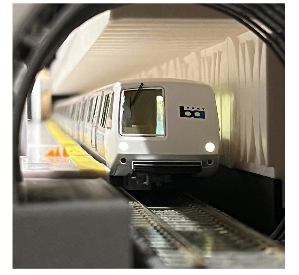miniature BART commuter train with white nose, two bright white lights, large front window and black windshield wiper in a modeled underground station scene.