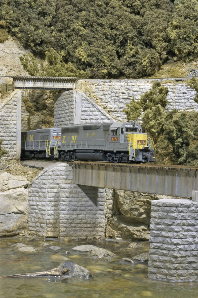 A model train crosses over a river bridge ion a model railroad layout
