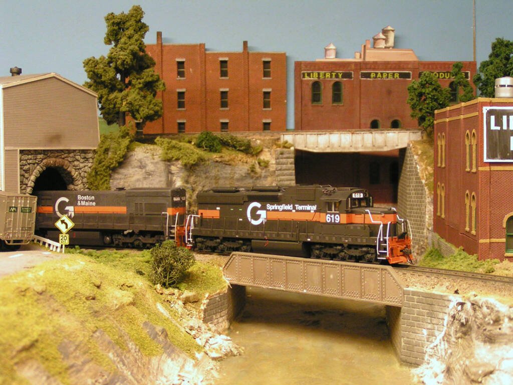 A model diesel locomotive leads a train across a bridge over a river on a model railroad layout