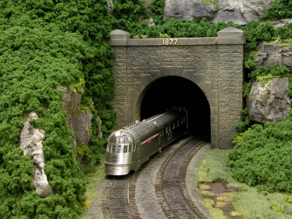 A silver streamlined passenger train is entering a forested tunnel on a model railroad layout