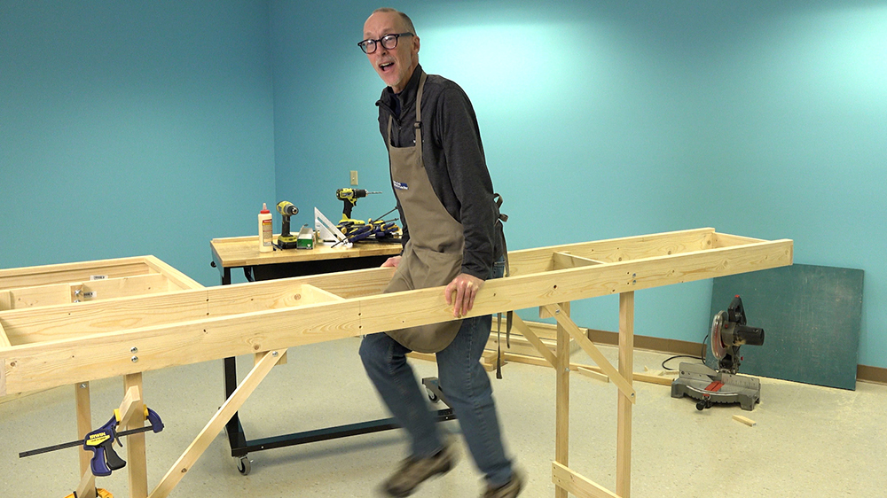 White male supporting himself with extended arms holding onto an open-frame construction made from bare wood parts and held up on small wood legs.