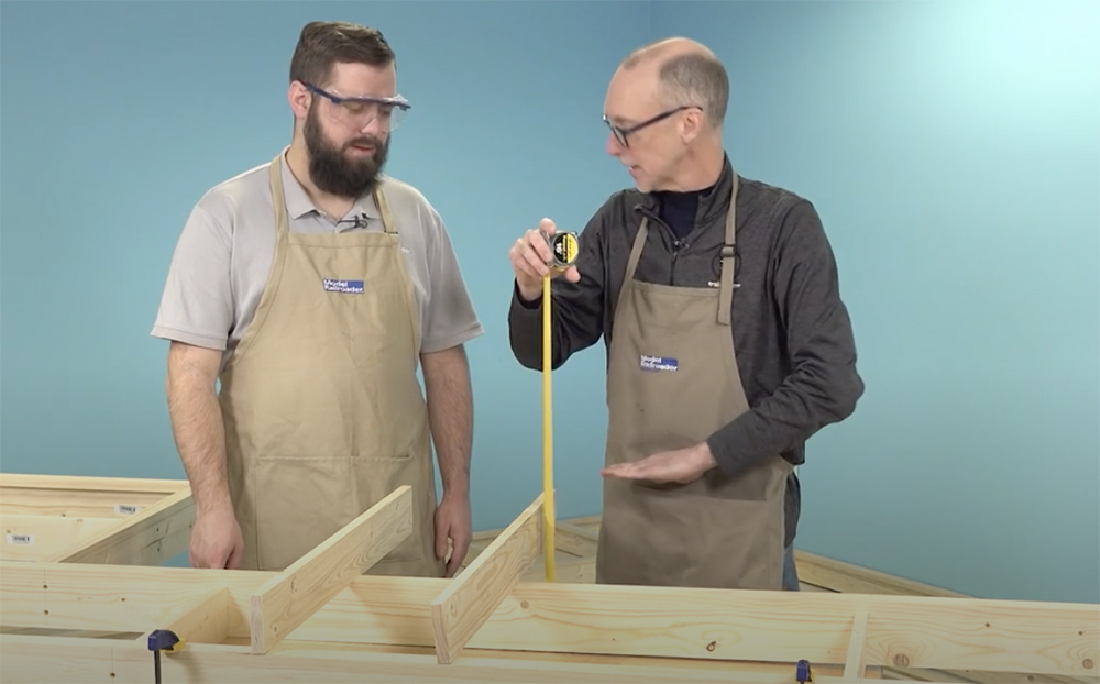Two white males, one with a full beard and one balding, hold a silver and yellow tape measure next to a bare wood frame with two boards resting on edge atop it.