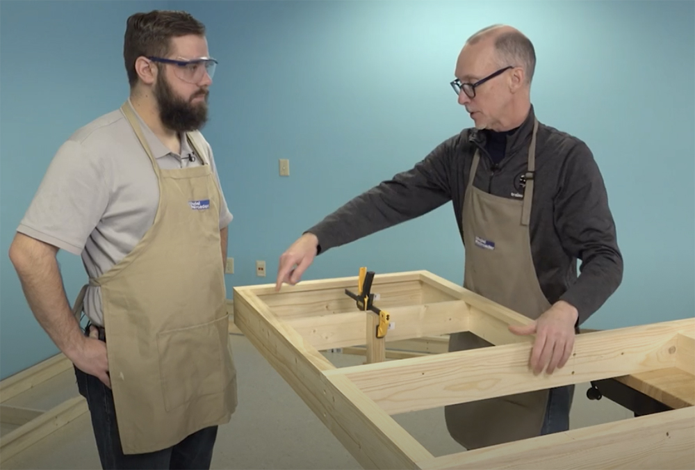 Two white males, one with a full beard and one with a balding head, stand at the end of a wood table frame with no top on it.