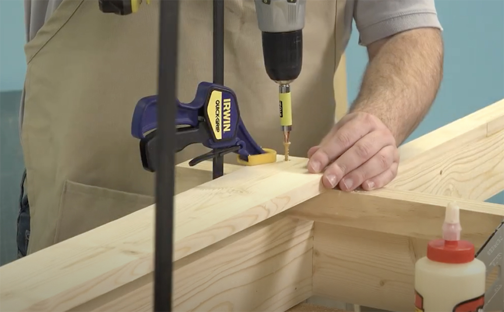Hands of a white, male carpenter holding a yellow deck screw in one hand and a green drill in the other as he drives the screw into tan-colored wood.
