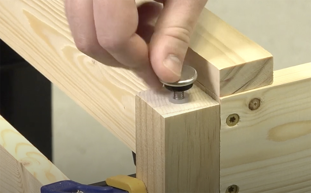 White male hand holding a silver metal disc with a threaded rod end over a 2 x 2 wood table leg.
