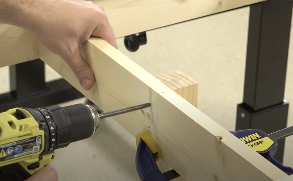 Yellow, black and silver drill being used to bore a hole through two tan pieces of lumber clamped together with black, blue, and yellow bar clamps.