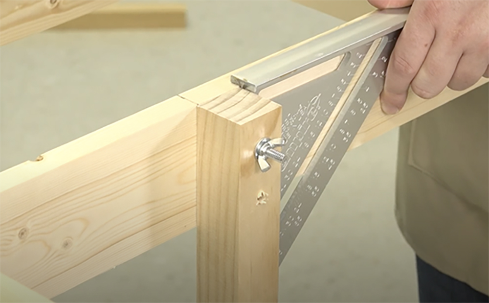 White male hand holding a silver triangular carpenter’s square next to a wood furniture leg attached with one silver bolt, washer, and wingnut to a bare wood table frame.