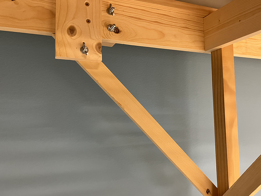Tan bare wood boards arranged as a table frame with a single square furniture leg that is braced by a narrow board placed at a 45-degree angle and held secured with silver bolts, washers, and screws.