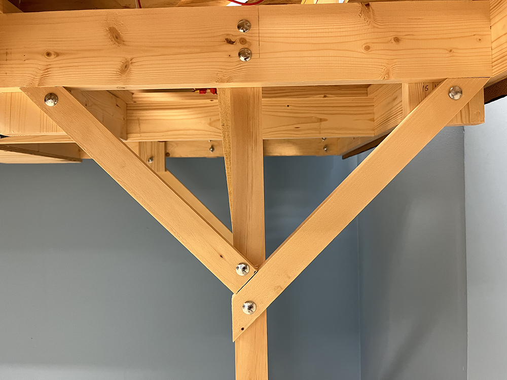 Wood square furniture leg with two braces set at opposite 45-degree angles where the leg connects to a bare wood table frame with all pieces attached using sliver bolts, washers, and nuts.