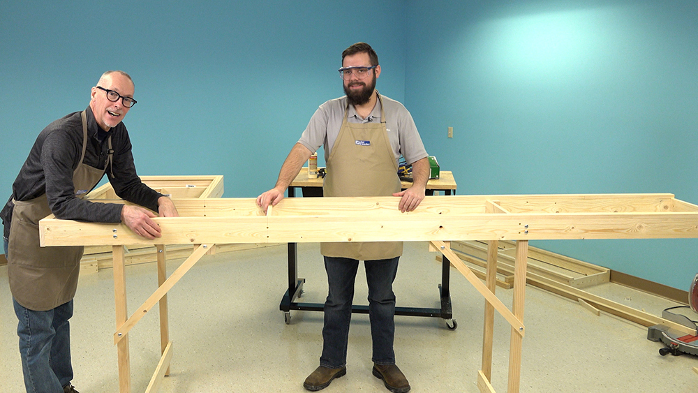 Two white males standing next to a rectangular wood frame supported by four square wood legs.