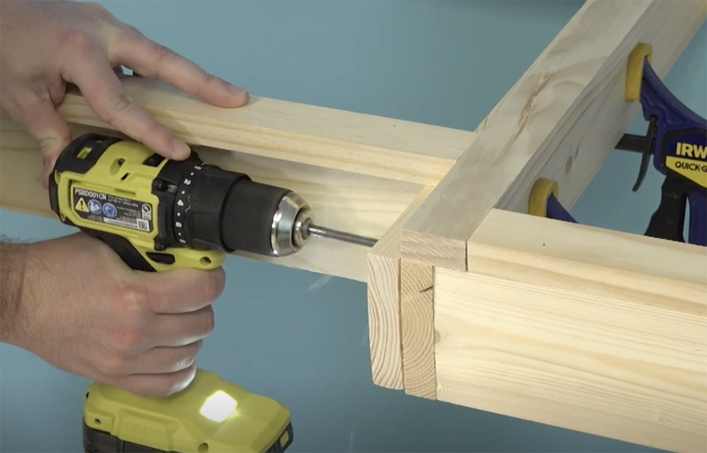 Yellow, black and silver drill being used to bore a hole in two bare wood rectangular frames held together by two black, blue, and yellow bar clamps.