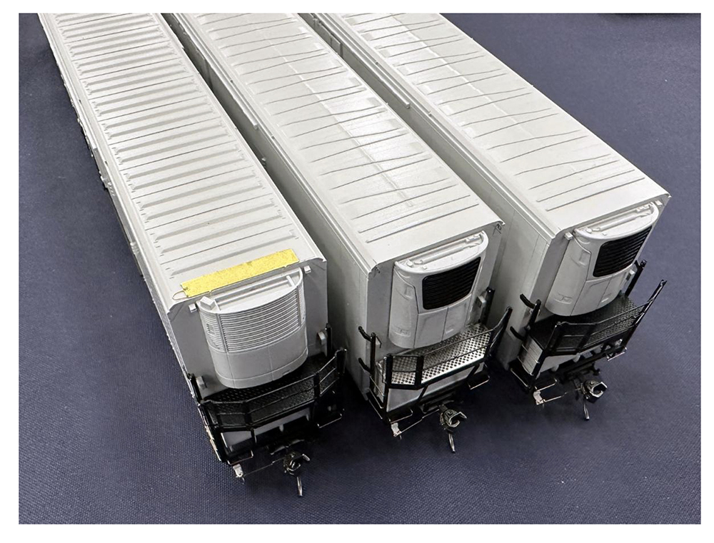 End view of three model train refrigerator cars in bare gray plastic and metal looking down on the ends with the refrigeration unit with the roof visible.