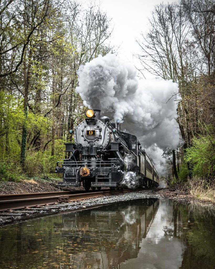 Black and gray steam locomotive with large smoke plume running in woods.