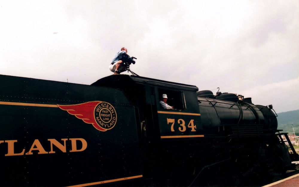 Cameraman sits on top of steam locomotive cab