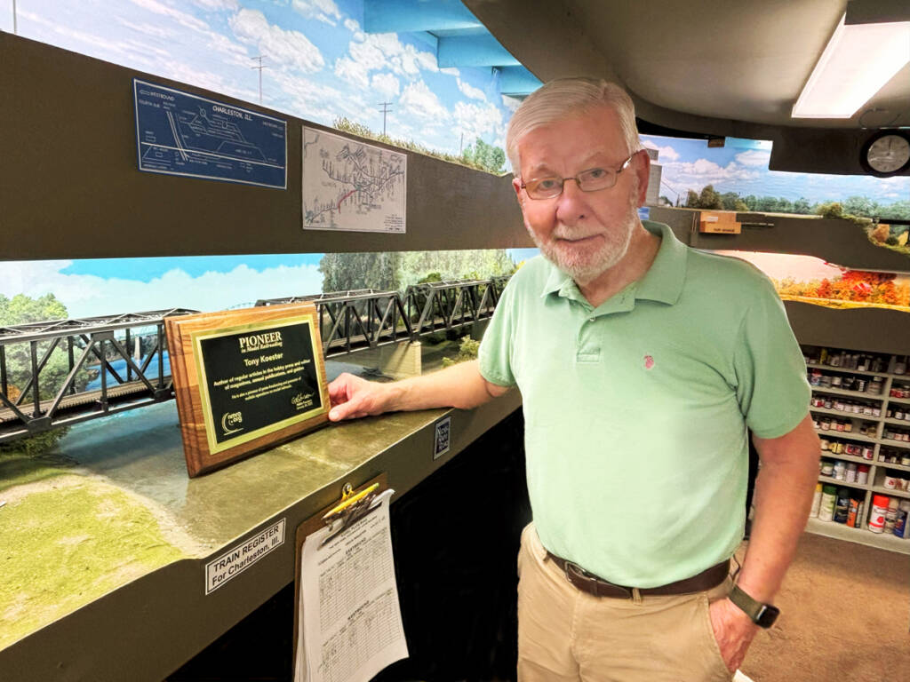 Color photo of man with gray hair and beard wearing glasses, green shirt, and khaki pants standing in front of an HO scale model railroad with an award.
