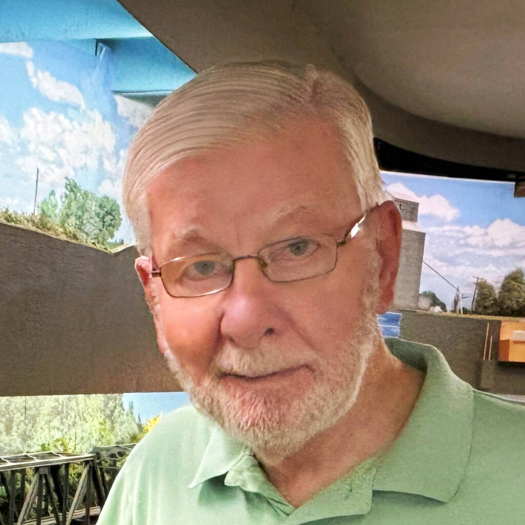 Color photo of man with gray hair and beard wearing glasses and green shirt standing in front of an HO scale model railroad.