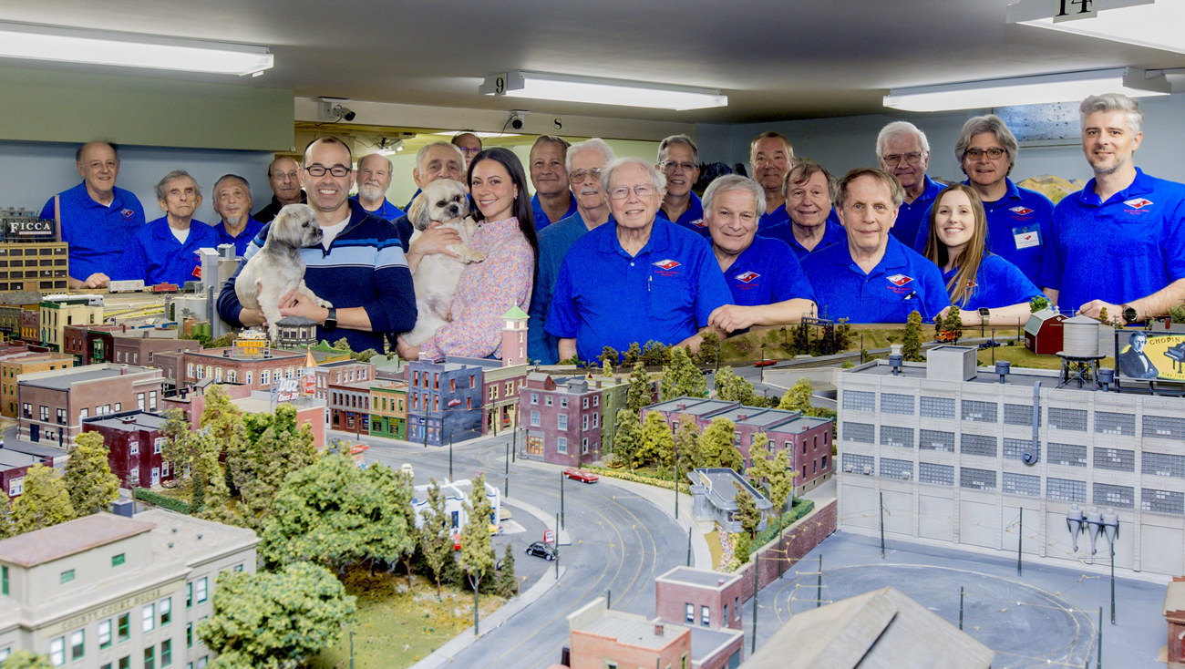 A group of people and two dogs stand behind a section of a large model railroad layout