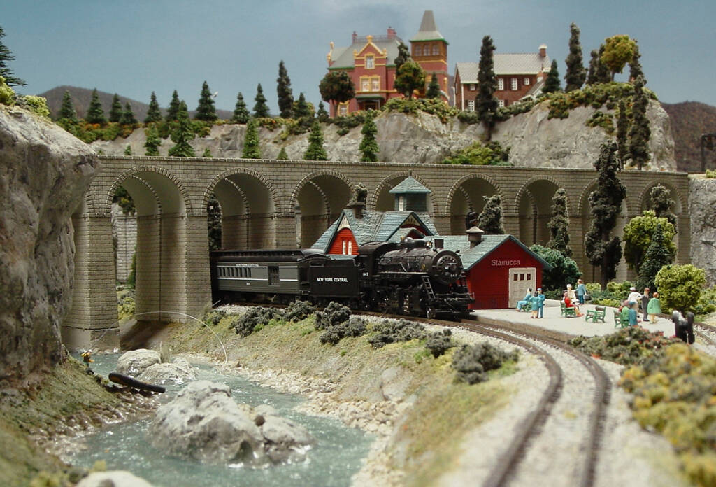 A model train pull up to a passemger station next to a river with a bridge above the train