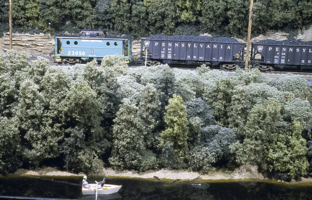A model train passes a small fishing boat on a river on a model railroad layout