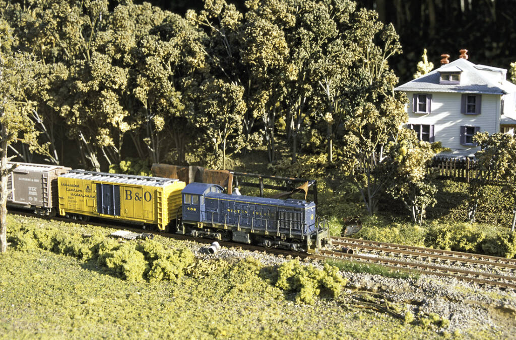 A blue diesel locomotive leads a train past a farm house on a model railroad layout