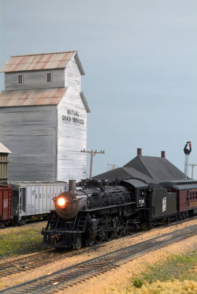 A model steam locomotive and tender pass a tall structure on a model railroad layout