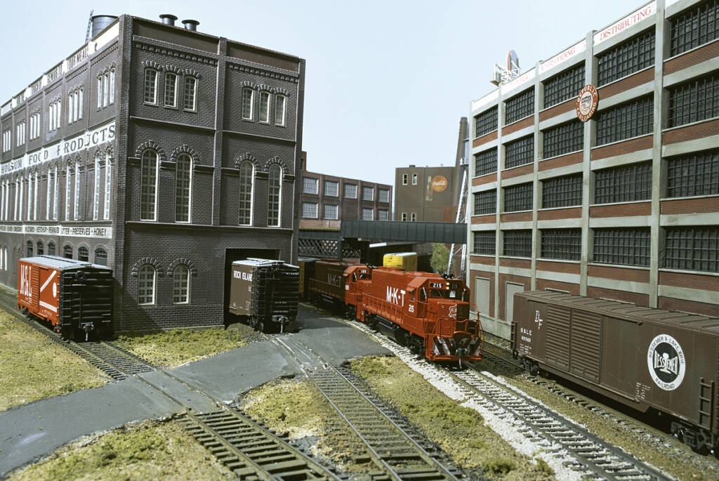 A red model locomotive leads a train at an industrial setting on a model railroad