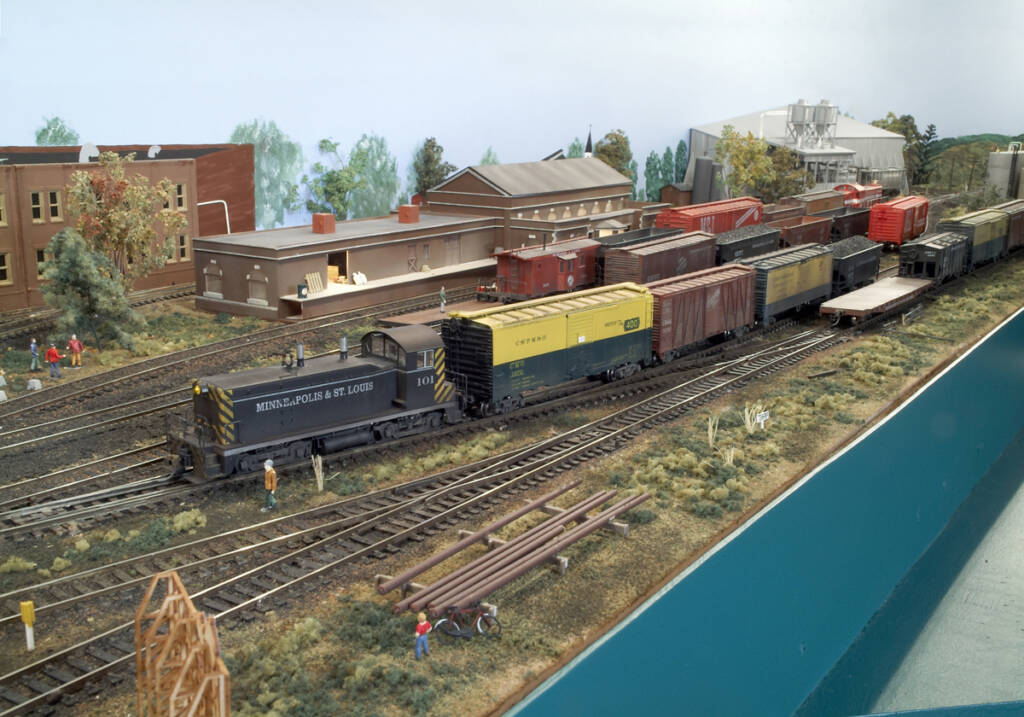 A model locomotive leads a freight train in a yard on a model railroad layout